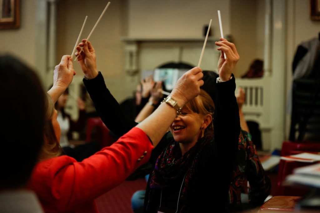 Two music teachers raise their arms while holding conducting batons, during a Music Teacher Mentoring session.