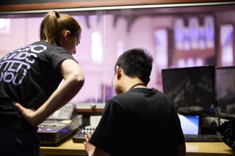 Two people are looking at a console with their backs to the camera. In the background is a concert hall