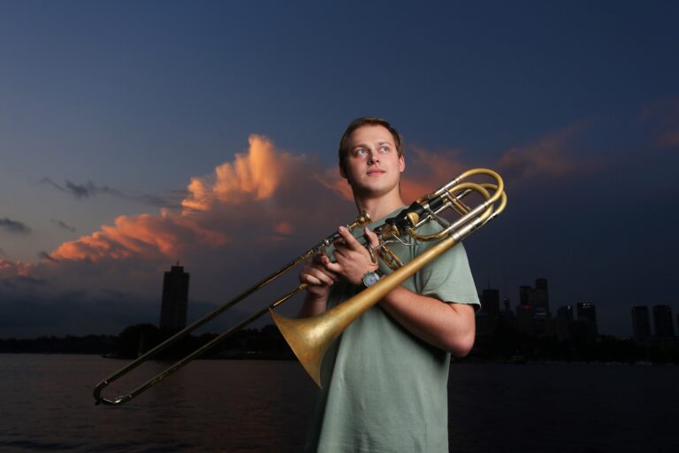 A young man holds a bass trombone. He is wearing a light green t shirt and behind him is a blue sky with pink and white clouds