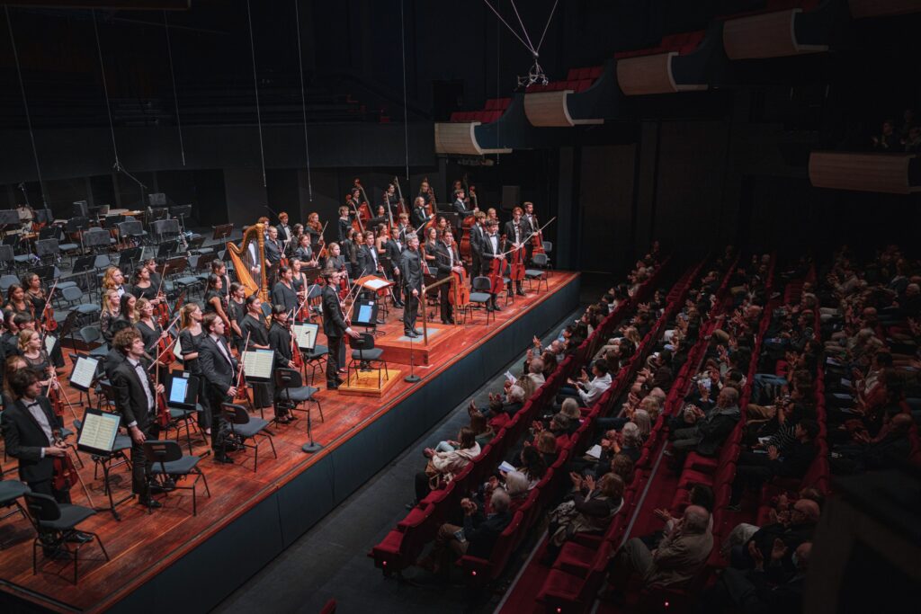 An orchestra of young musicians stand on stage for applause.