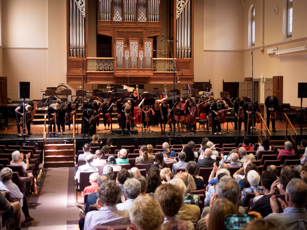 Members of an orchestra bow on stage after a performance, while the audience applauds.