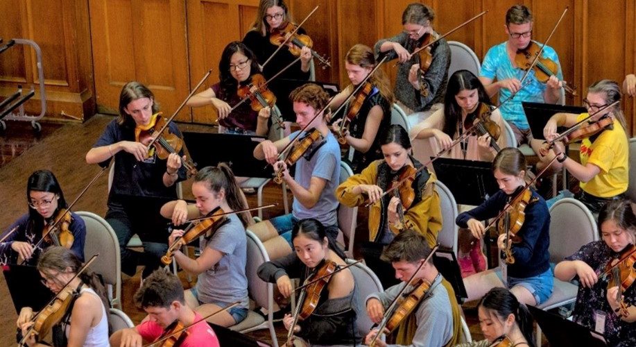 Reflections on Words About Music. An overhead view of a string section of an orchestra rehearses in a wood panelled room.