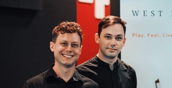 Two men stand side by side facing toward. They are smiling and wearing black shirts.