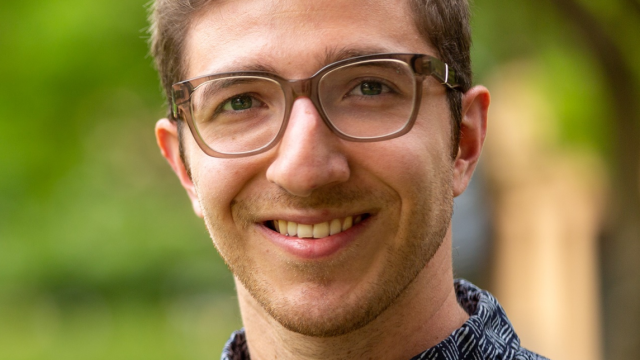 AYO Words About Music alumnus Adam Weitzer smiles at the camera. He is wearing a blue patterned button-up shirt and glasses.