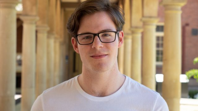 AYO Composition alumnus smiles and faces the camera. He is wearing a white tshirt and glasses.