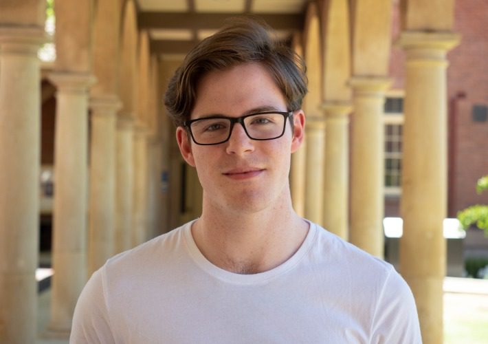 AYO Composition alumnus smiles and faces the camera. He is wearing a white tshirt and glasses.