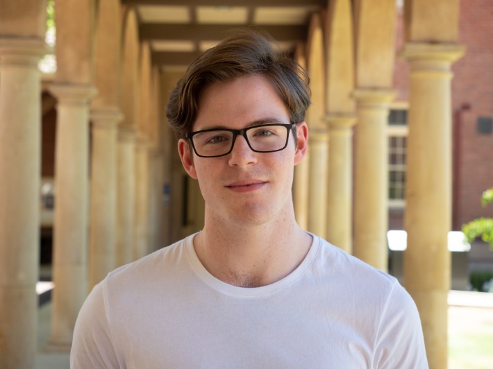 AYO Composition program alumnus Alexander Voltz smiles at the camera. He is wearing black-rimmed glasses and a white t-shirt.