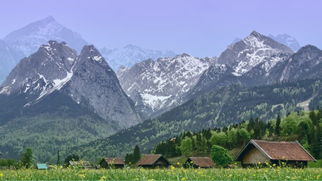 A grass field with houses and large mountains in the distance. The Image has a blue background.