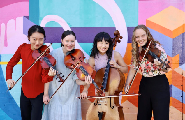 A string quartet pose with their instruments in front of a colourful wall mural. They are all smiling and looking directly at the camera.