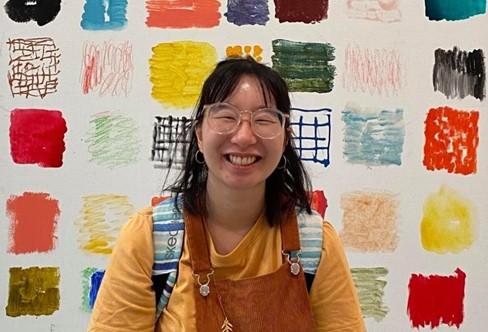 Ashleigh Ho 2021 AYO Words About Music Participant smiles at the camera against a background of brightly coloured squares. She is wearing a canary yellow shirt and is wearing a backpack and glasses.