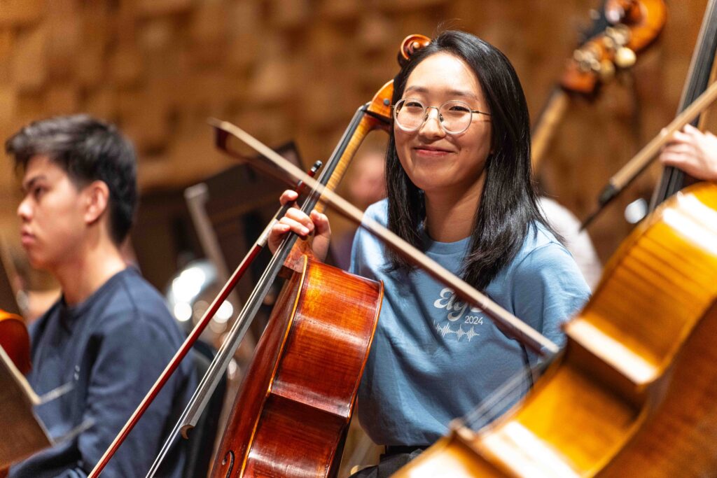 Girl in blue AYO t shirt smiles at the camera holding a cello