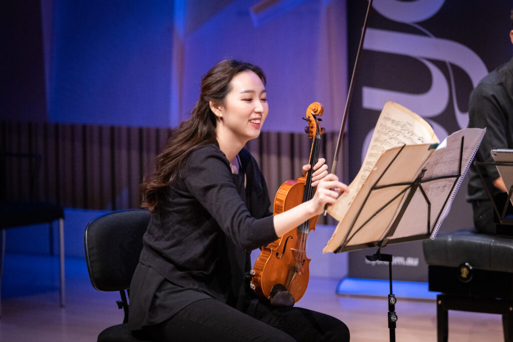 A violinist smiles as she leans forward and makes a note on her sheet music.