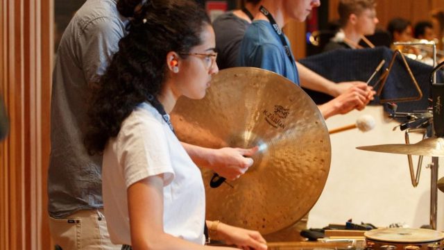 AYO Percussionists pictured from the side facing right standing in front of percussion instruments.