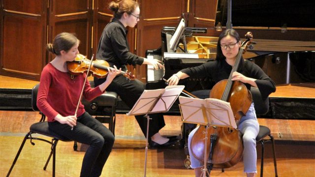 A piano trio rehearses on stage in an auditorium.