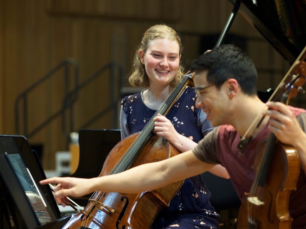 A young woman with short blonde hair in a blue dress laughs with a young man with short black hair and glasses during a rehearsal. They are both sitting down, she holds a cello and he holds a viola.