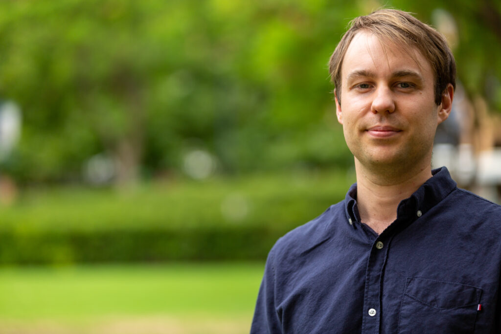 Composer and Words About Music participant Christopher Healey smiles at the camera. He is outside and surrounded by lush green plants, 