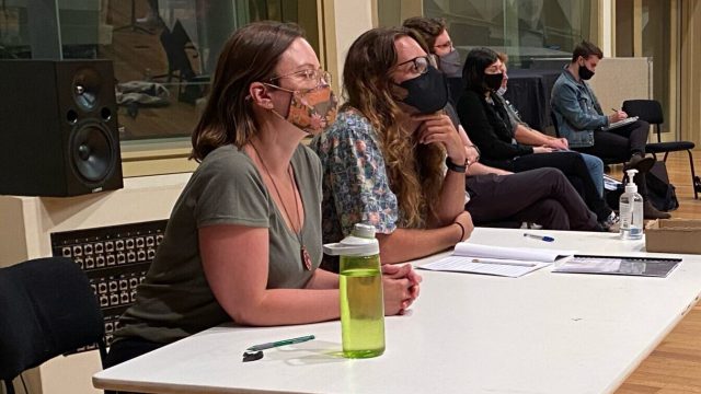 A row of people sit at a table facing right of frame. They are wearing masks.