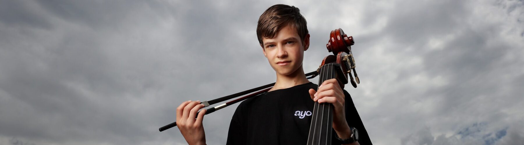 Young man looks at camera in front of a cloudy sky. He is holding a double bass