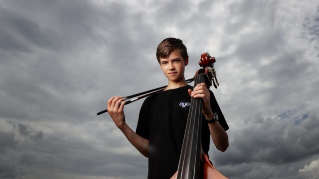 Young man looks at camera in front of a cloudy sky. He is holding a double bass