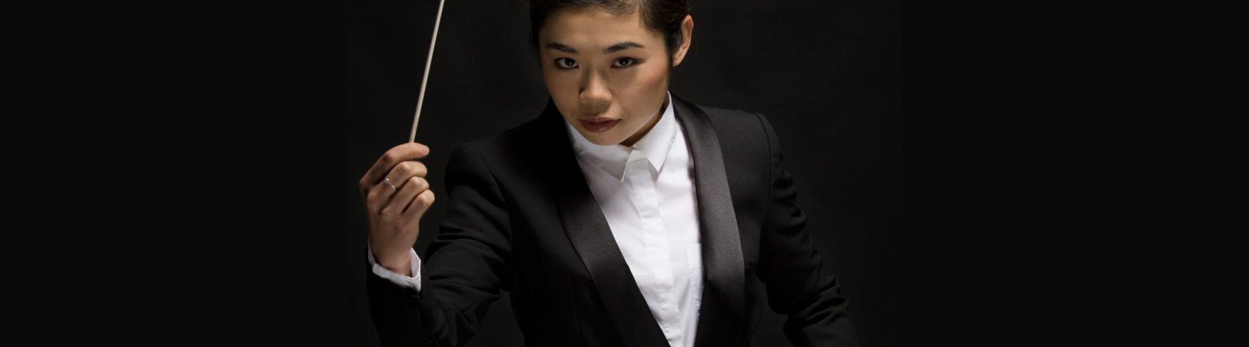 Conductor Elim Chan raises her conductor's baton in one hand. She is wearing a white shirt and tailored black suit jacket.