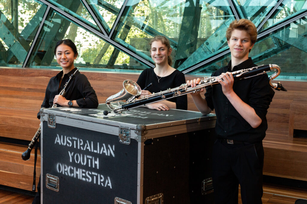 AYO musicians Ruby Khuu, Kara Thorpe and Adam Clennar smile at the camera as they hold their auxiliary instruments. They are dressed in concert blacks.