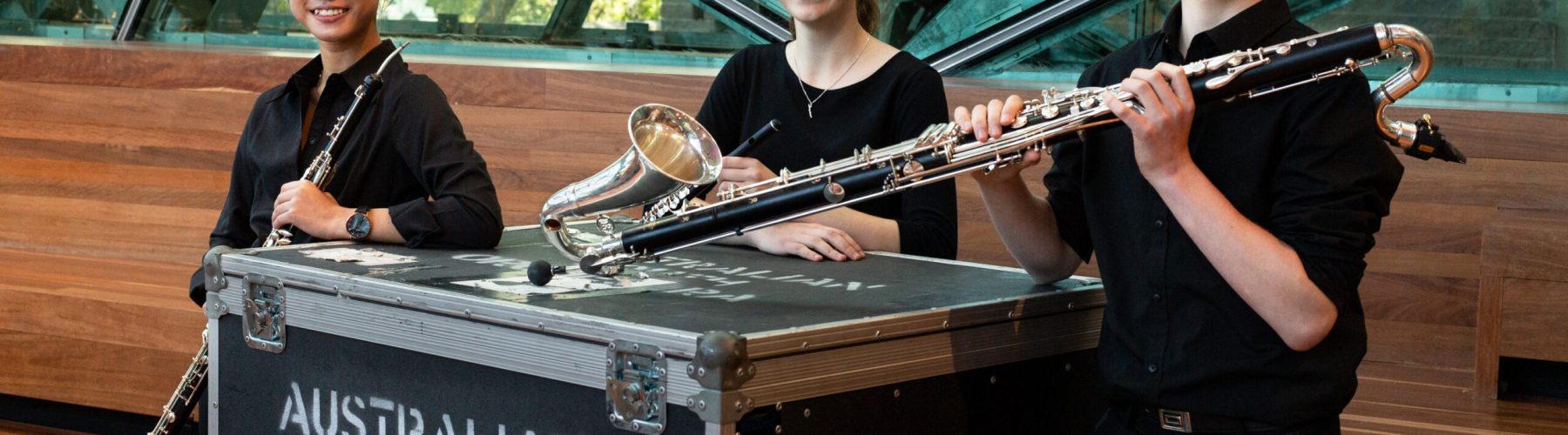 AYO musicians Ruby Khuu, Kara Thorpe and Adam Clennar smile at the camera as they hold their auxiliary instruments. They are dressed in concert blacks.