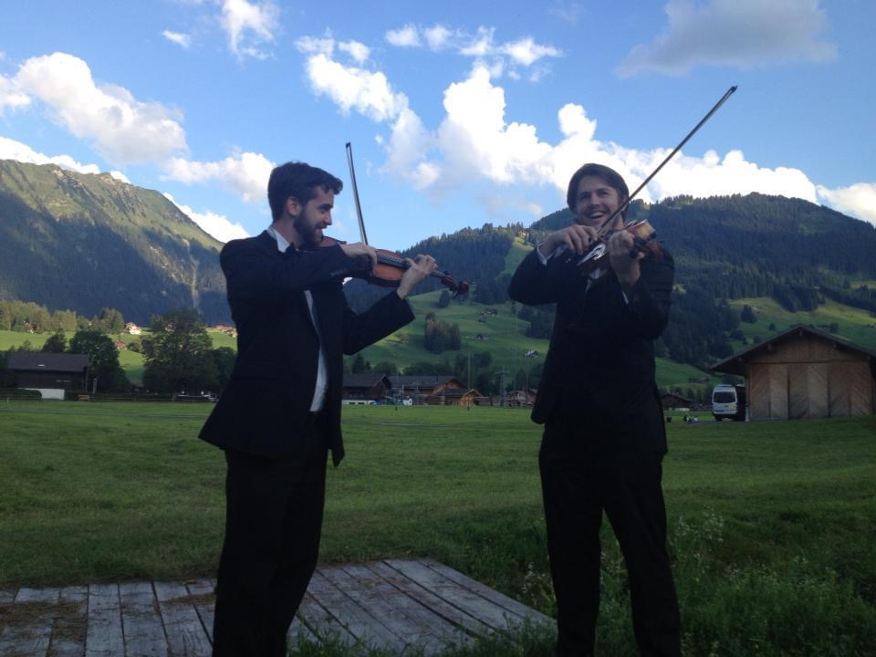 A violist and violinist play their instruments outside with Swiss mountains in the background. 