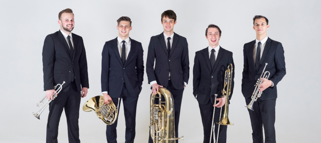 Five young men dressed in black suits smile at the camera. They are standing in front of a grey-white background and are each holding a brass instrument.