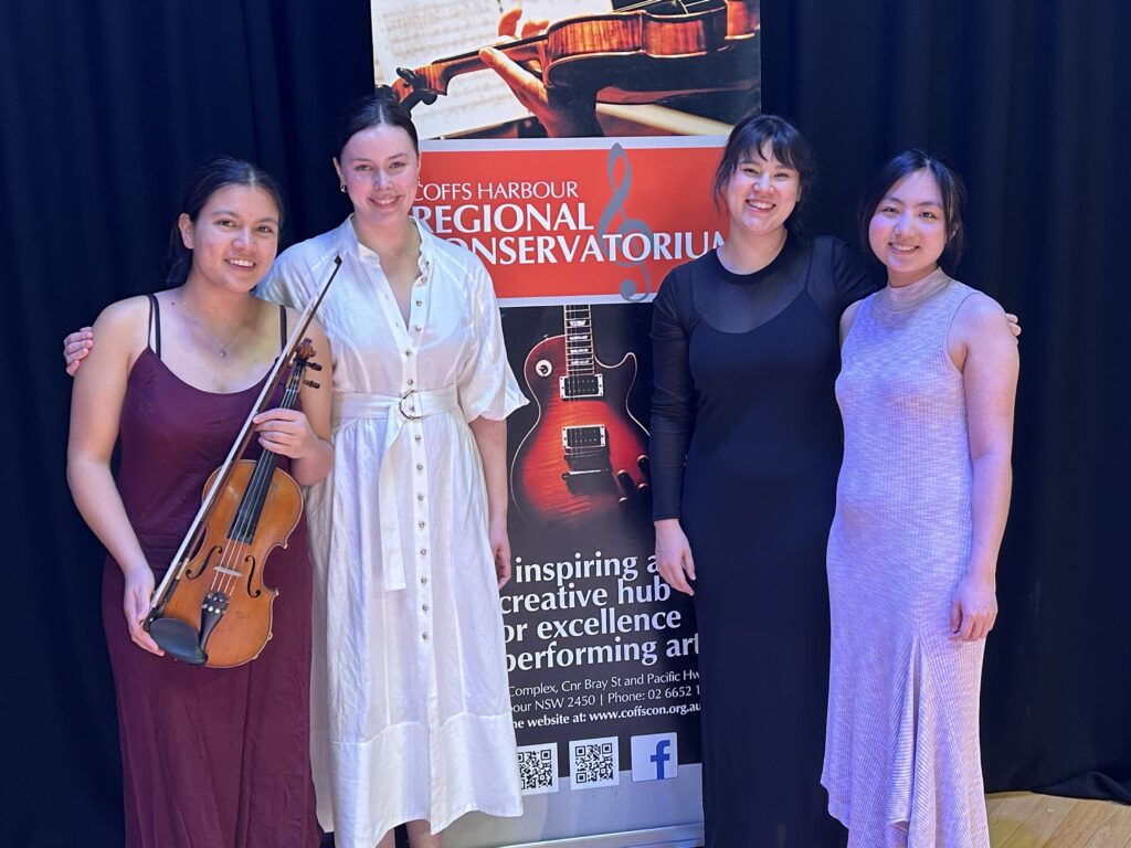 Amiti String quartet smile on stage while standing in front of a Coffs Harbour Regional Conservatorium banner.