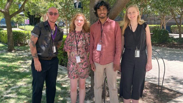 Four people stand outside under a tree.