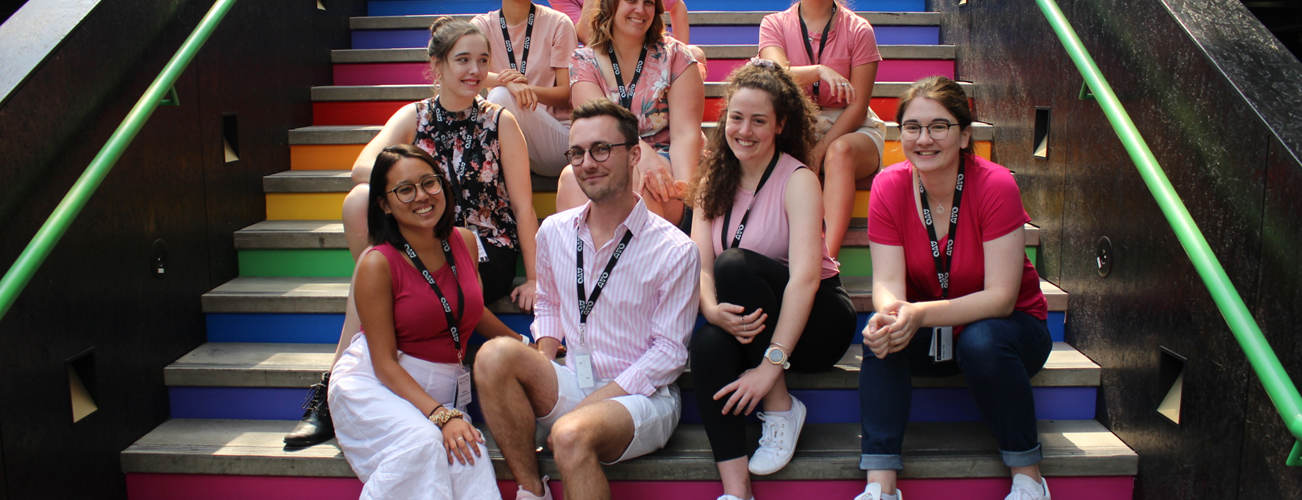 The Orchestral Management cohort from AYO National Music Camp 2020 sit in a group on a colourful flight of outdoor stairs. They are smiling at the camera.