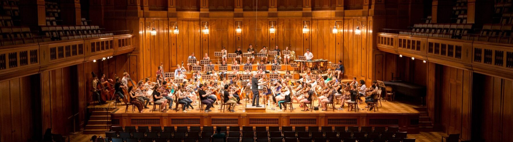 An orchestra rehearses in a concert hall with ornate chandeliers hanging from the ceiling.