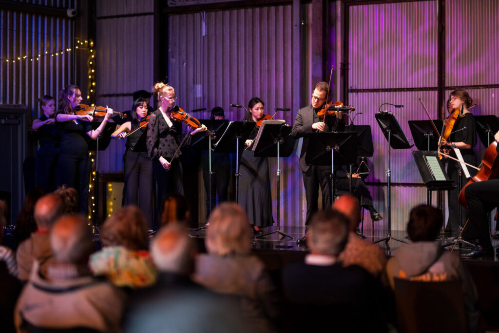 A string orchetsra performs in a venue with corrugated iron walls. 