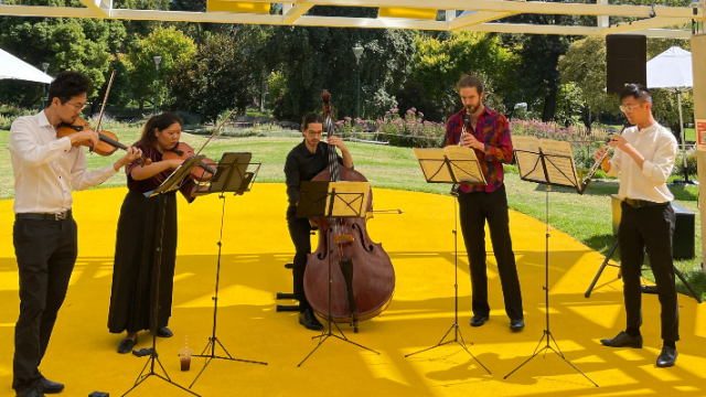 An ensemble of 5 musicians standing under a metal pavilion playing instruments. They are outdoors on a sunny day and the floor is bright yellow.