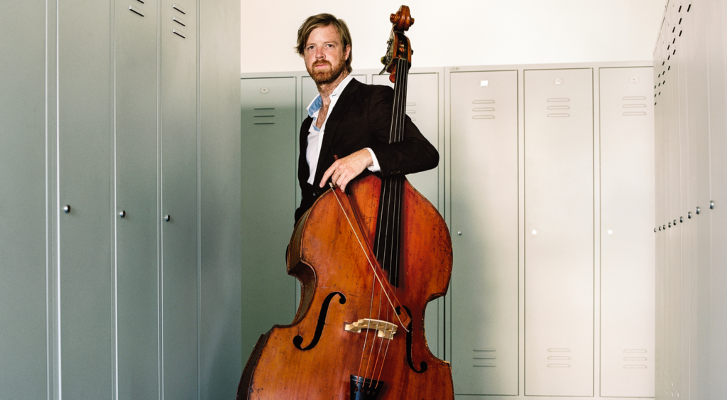 AYO alumnus and double bassist Matthew McDonald stands with his instrument. He is wearing a black suit jacket.