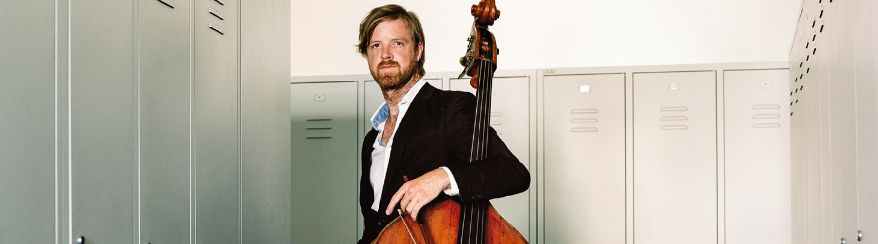 AYO alumnus and double bassist Matthew McDonald stands with his instrument. He is wearing a black suit jacket.