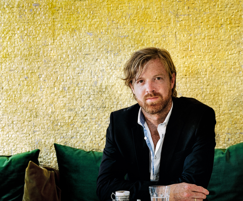 AYO alumnus and double bassist Matthew McDonald smiles at the camera. He is sitting down and behind him is a textured yellow wall. 