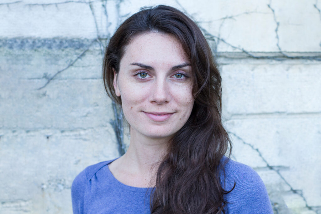 Composer Melody Eötvös smiles at the camera. She is wearing a blue top and has long brown hair.