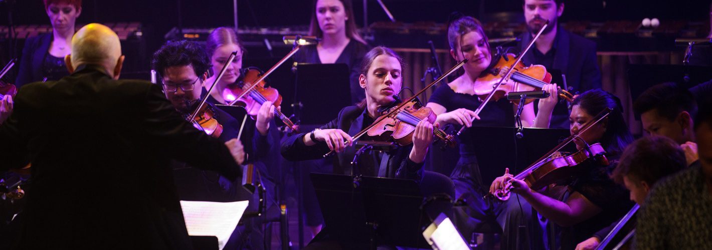 String players perform in an ensemble. they are facing a conductor, pictured left and are lit by purple stage light.