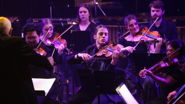 String players perform in an ensemble. they are facing a conductor, pictured left and are lit by purple stage light.