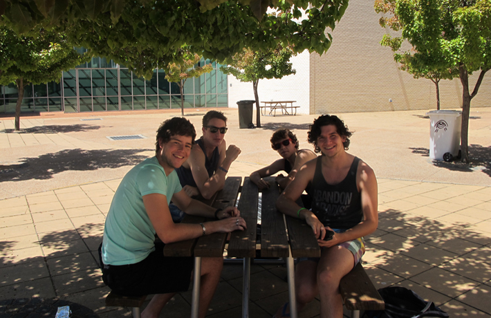A group of young musicians sit around a picnic table on a sunny day.