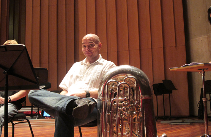 A man smiles at the camera while sitting on a chair, a tuba on the ground next to him.