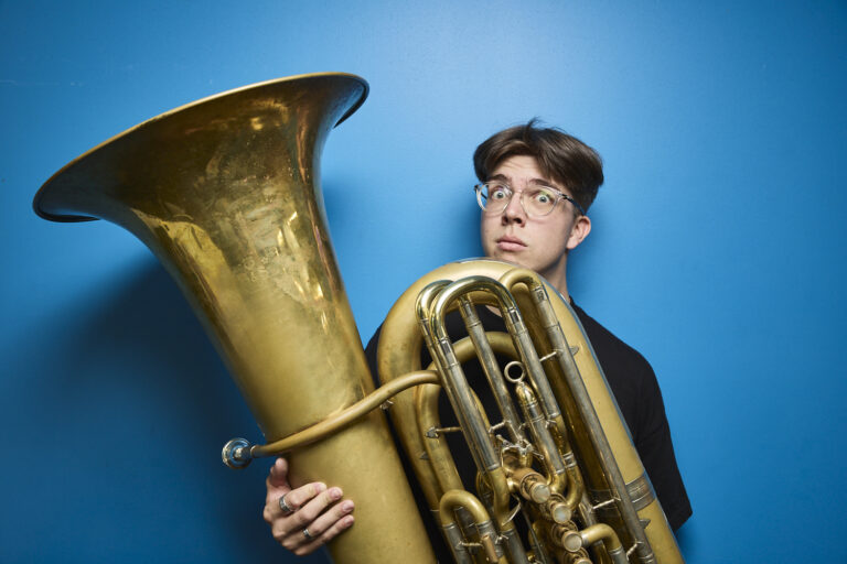 A young man wearing glasses holds a tuba in front of himself in front of a plain blue background. His eyebrows are raised comically.