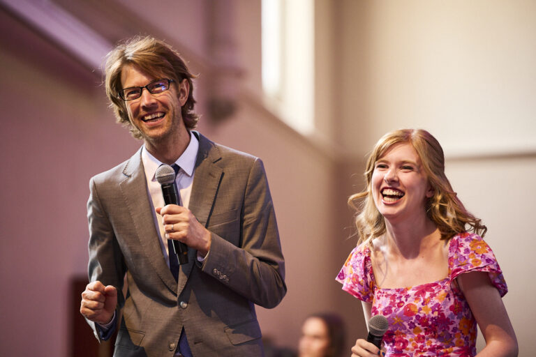 A man wearing a grey suit and a young woman wearing a pink floral dress are holding microphones and laughing