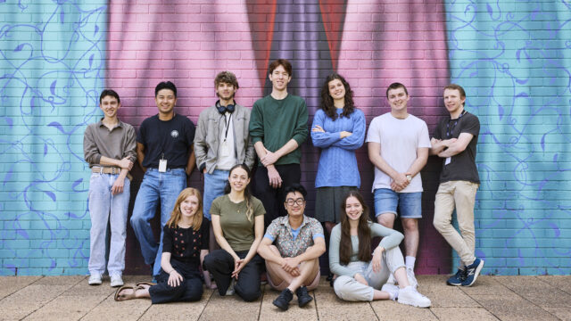 A group of eleven young people dressed in casual clothes stand in front of a brink wall with a painted mural.