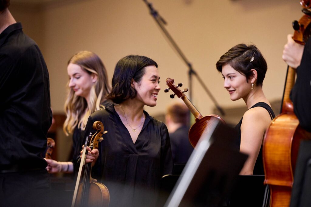 Two musicians of the Australian Youth Orchestra smile at each other during AYO National Music Camp. They are holding violas and wearing concert blacks. Credit Claudio Raschella 2023