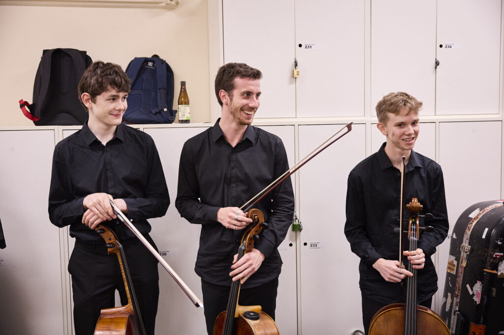 Three young cellists stand backstage dressed in concert blacks.