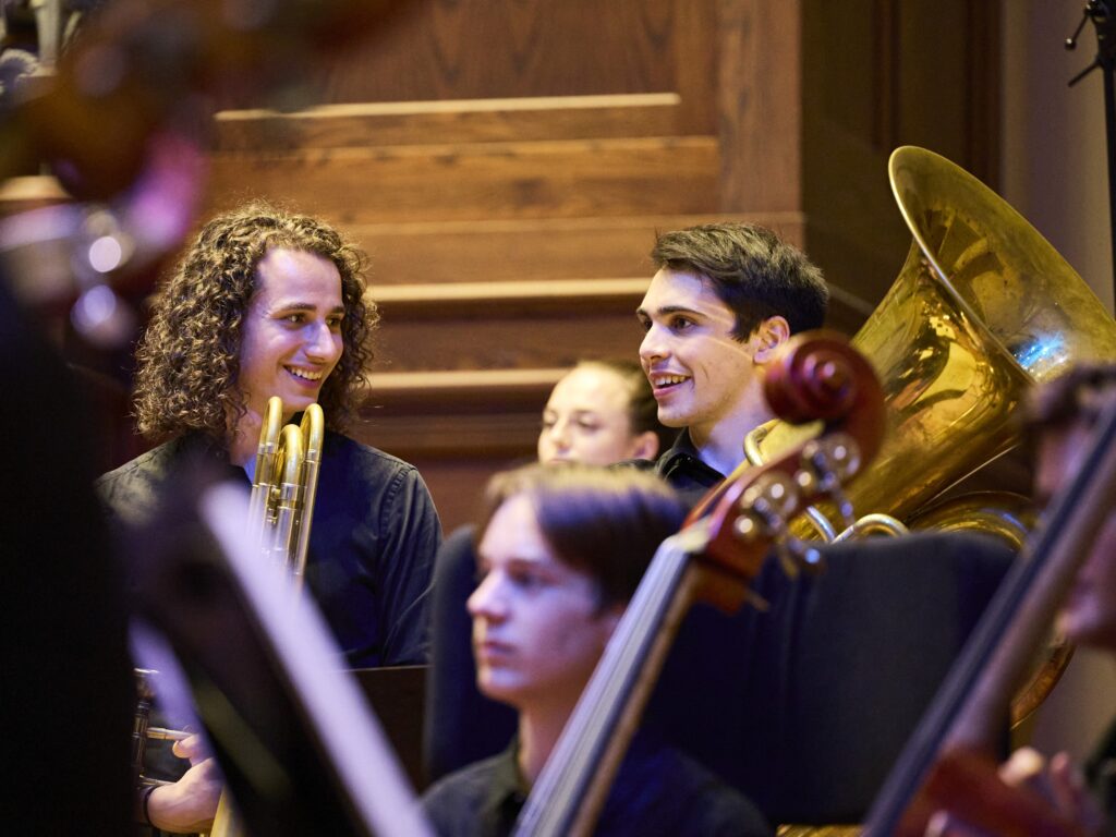 A trombone player and tuba player laugh together while standing for applause after a performance.