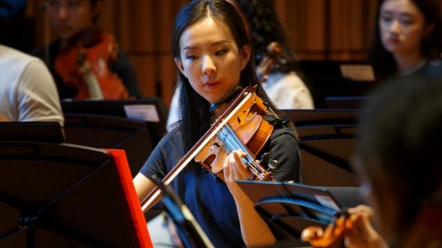 A female violinist sits in front of a music stand whilst practicing. She has long black hair and is wearing a dark tshirt.