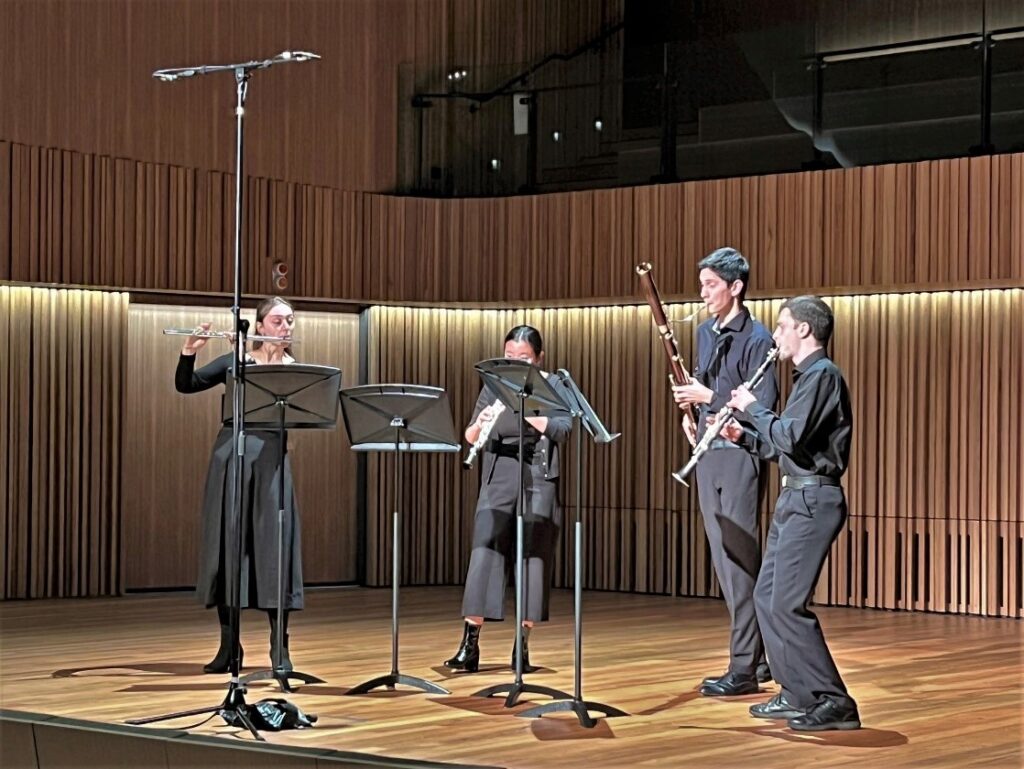 Four woodwind musicians dressed in concert blacks perform in an auditorium.